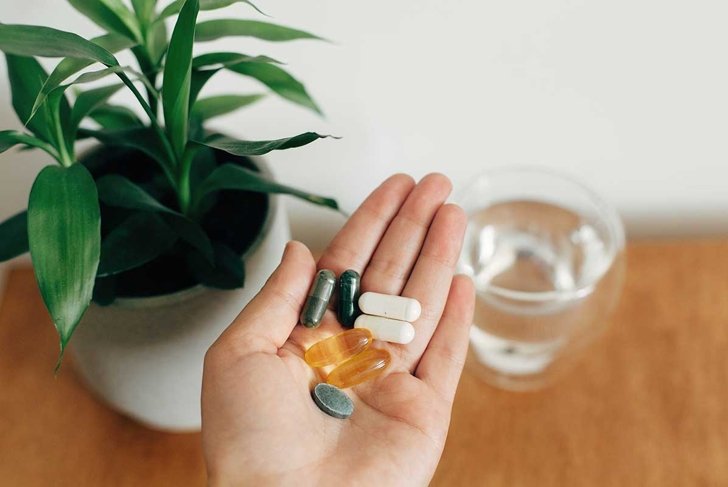 Hand holding omega 3, spirulina, chlorophyll,magnesium capsules above glass of water on wooden table. Morning vitamin pills. Dietary supplements. Health support. Biologically active additives