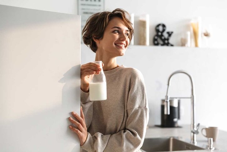 Happy brunette woman drinking milk and looking away while standing in kitchen