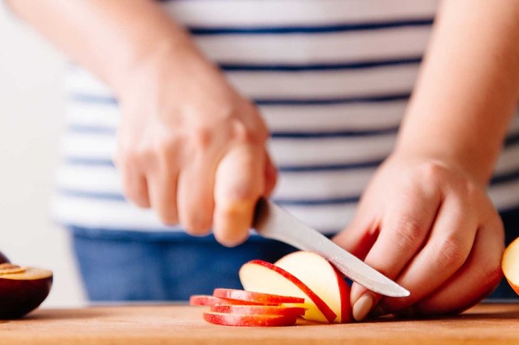 Woman cooking and cutting apple on wooden cutting board. Preparing healthy food. Hands holding knife and slicing fruits on table in kitchen