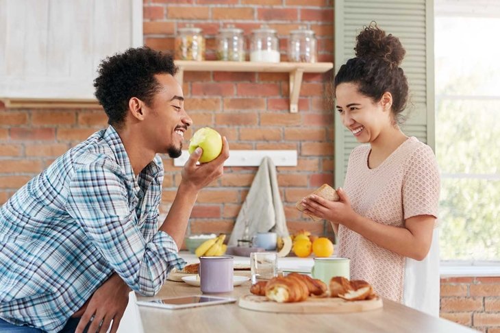 Newlyweds spend time together, speak pleasantly at kitchen, have good relationships. Smiling pretty brunette mixed race woman looks at husband who eats green apple and waits for delicious supper