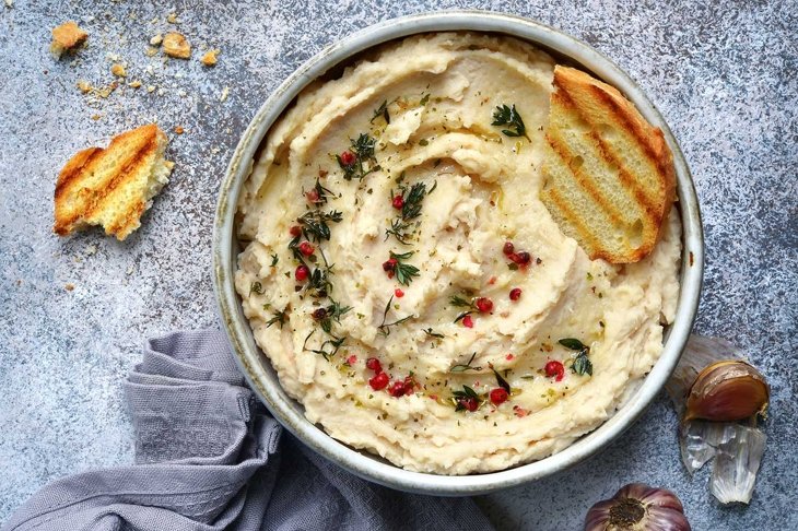 White bean hummus with baked garlic and dried herbs in a bowl over grey slate, stone or concrete background.Top view with copy space.